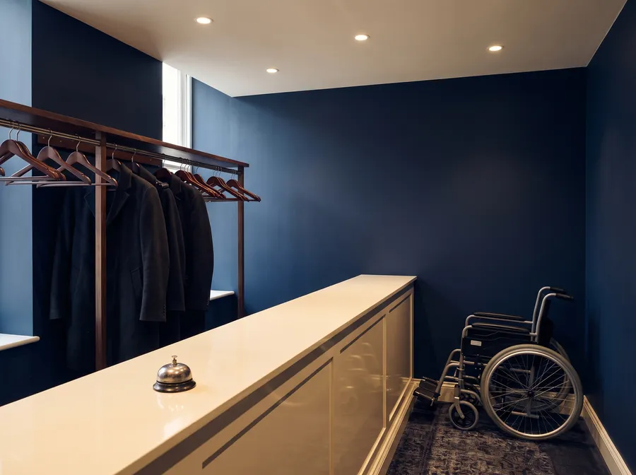 Scène générée : un vestiaire aux murs bleu marine, des manteaux sur des cintres, un comptoir crème avec une sonnette et un fauteuil roulant plié.
