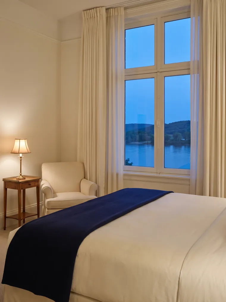Scène générée : une chambre crème au crépuscule, une couverture bleu marine sur le lit, un fauteuil et une haute fenêtre ouverte sur un fleuve très large.